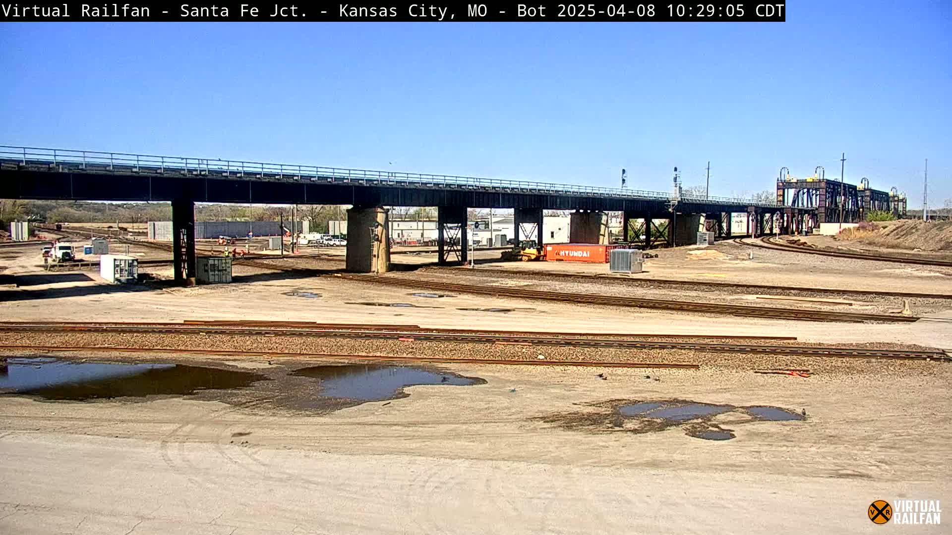 A mostly sunny day reveals a rail yard with a large bridge overhead, some construction equipment, and several puddles of water.