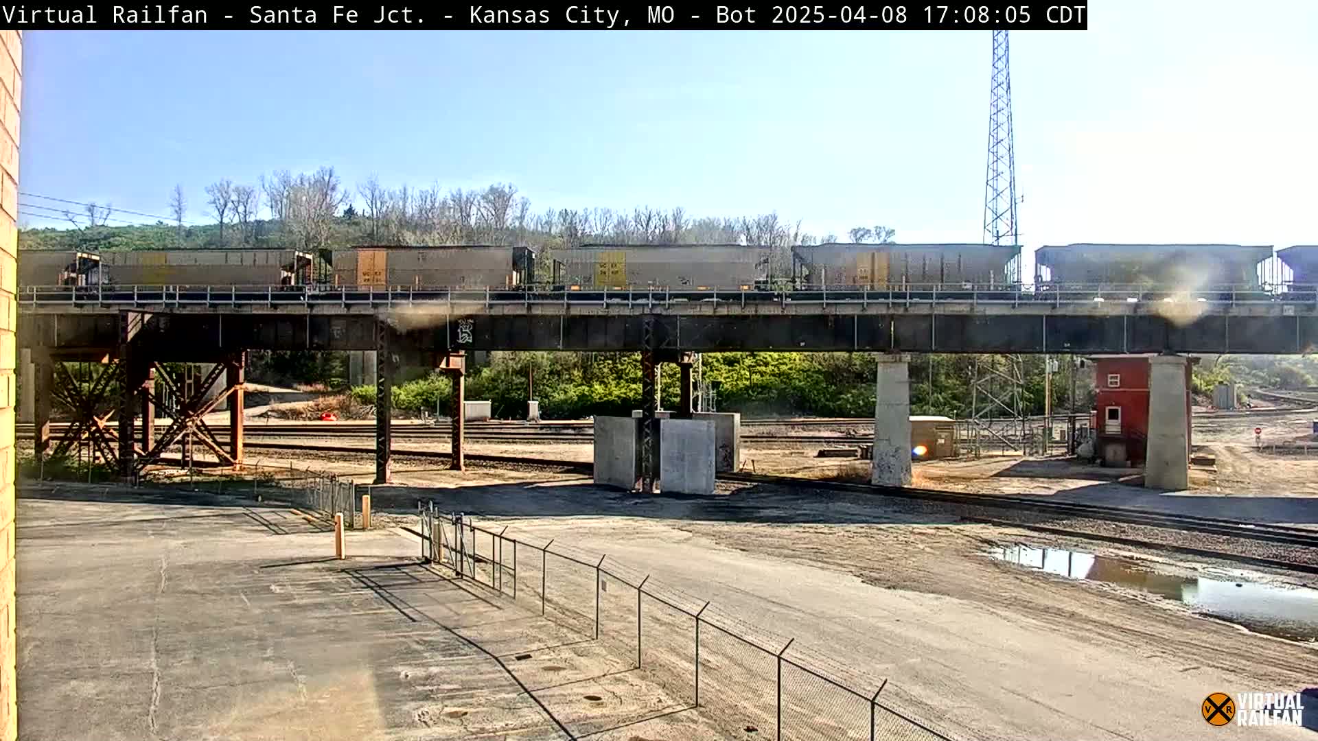 A freight train crosses a railway bridge under a mostly sunny sky, with a rail yard visible below.