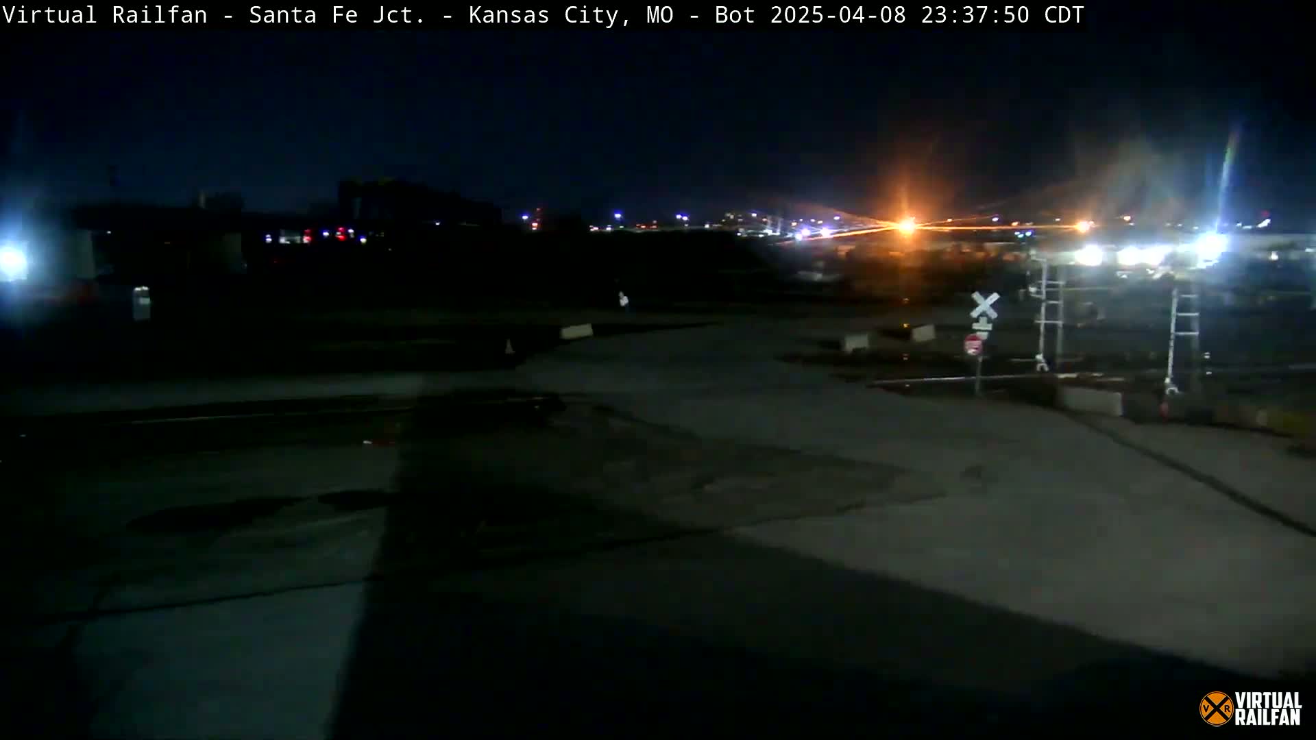 A nighttime view of a dark, mostly empty railroad crossing with a few lights visible in the distance.