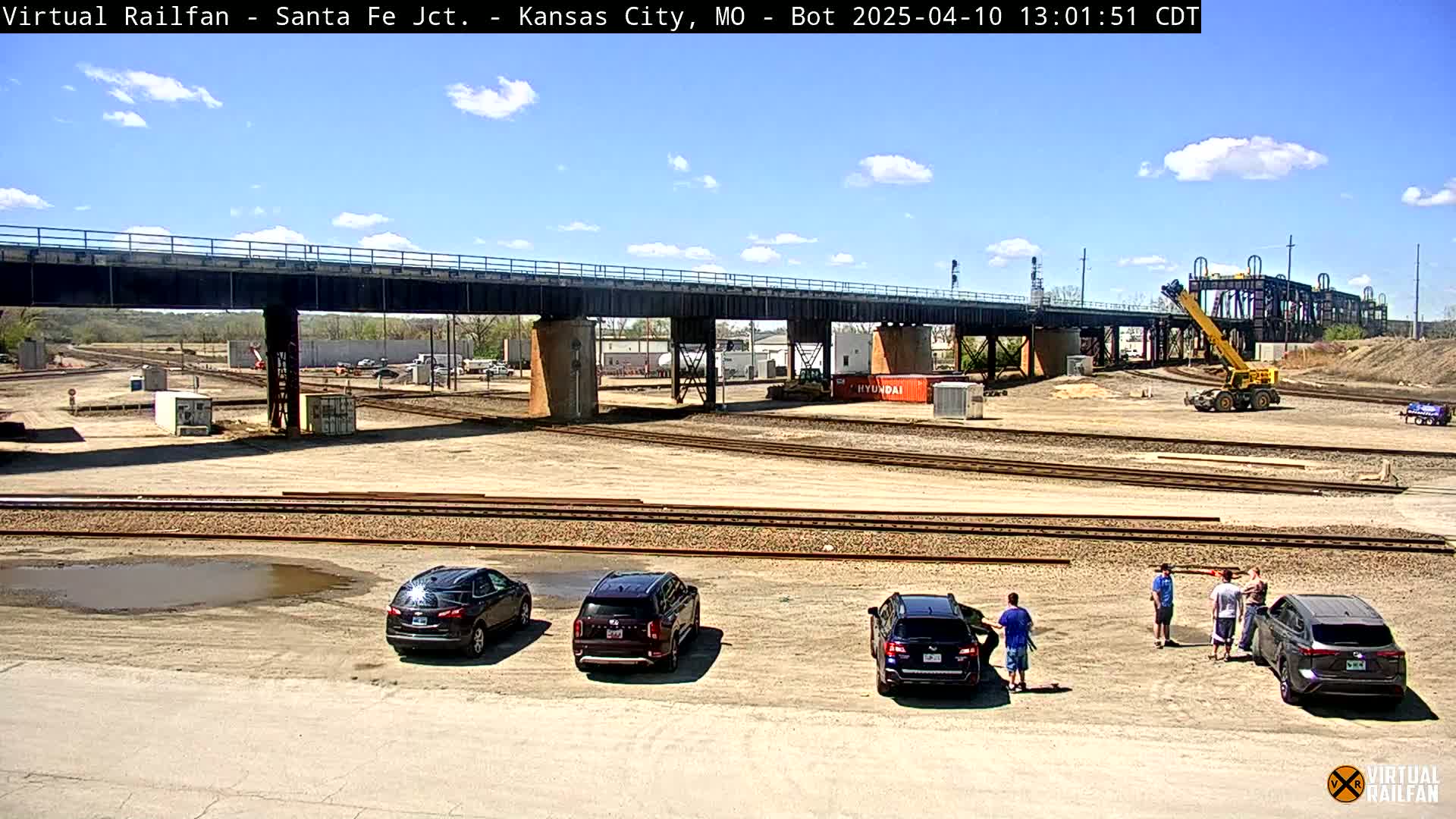 A sunny day shows a railway yard with several cars parked near a large bridge and a crane, with people standing near the vehicles.