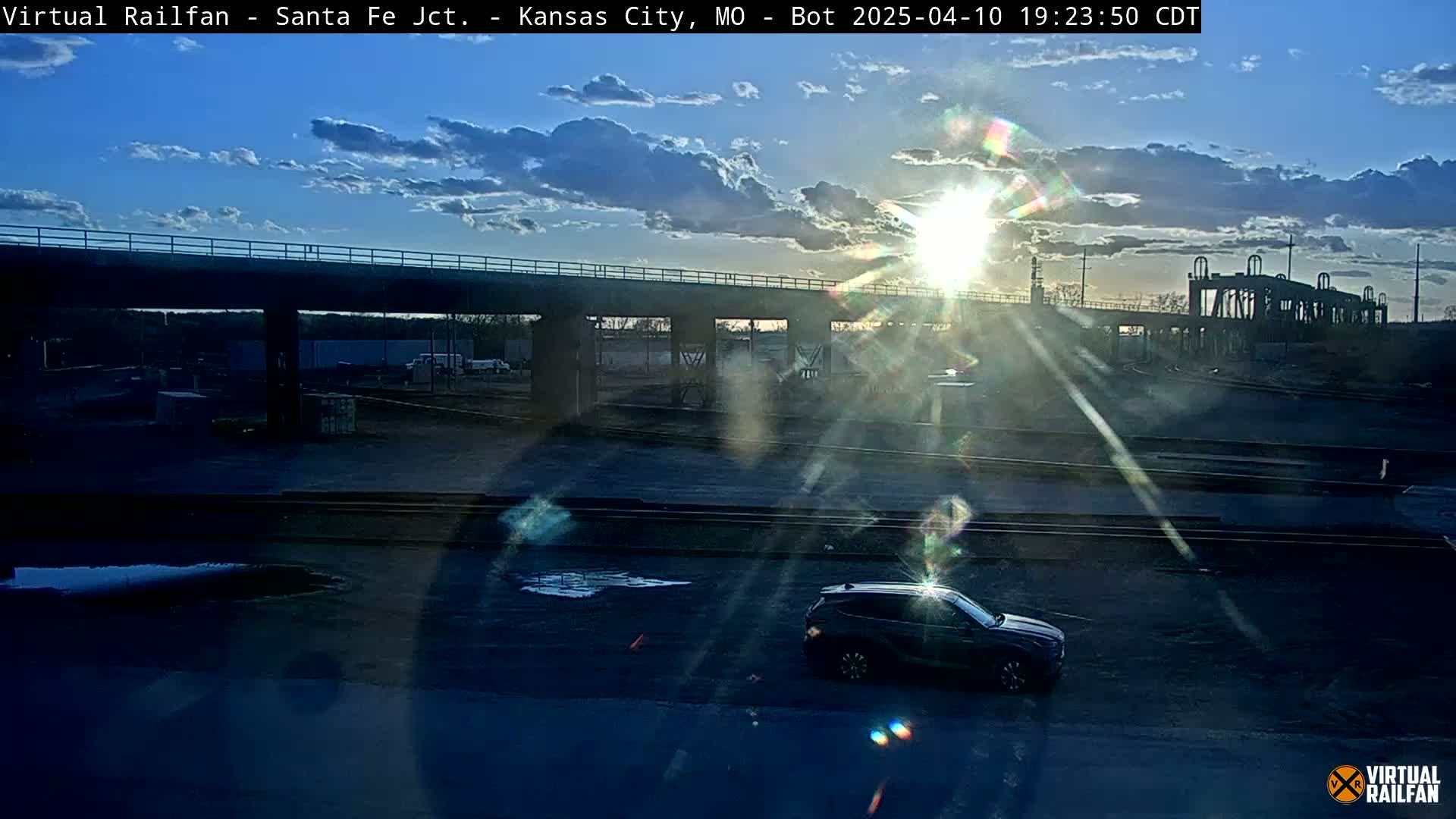 A dark-colored SUV is parked in a lot near a railway underpass during a partly cloudy sunset.