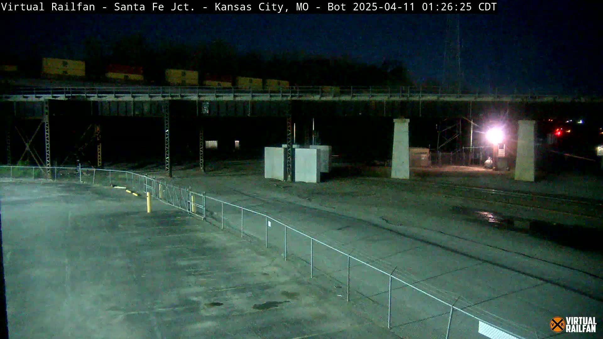 A nighttime view of a deserted rail yard under a dark sky, showing a metal structure and several large concrete blocks.