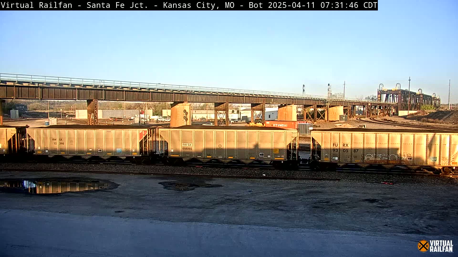 Several tan freight cars sit on railroad tracks under a bridge on a sunny day.