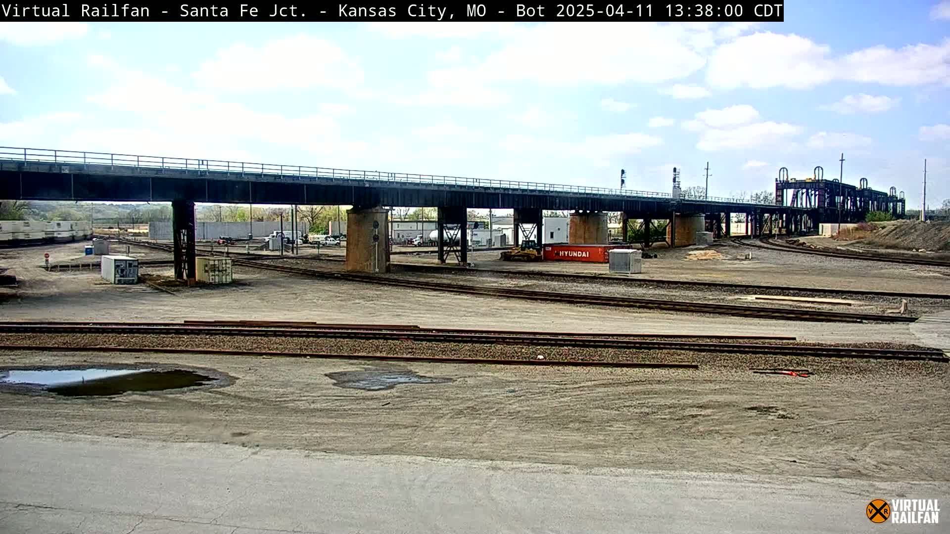 A mostly sunny day reveals a railway yard with multiple tracks, a large bridge overhead, some construction equipment, and a few shipping containers under the bridge.