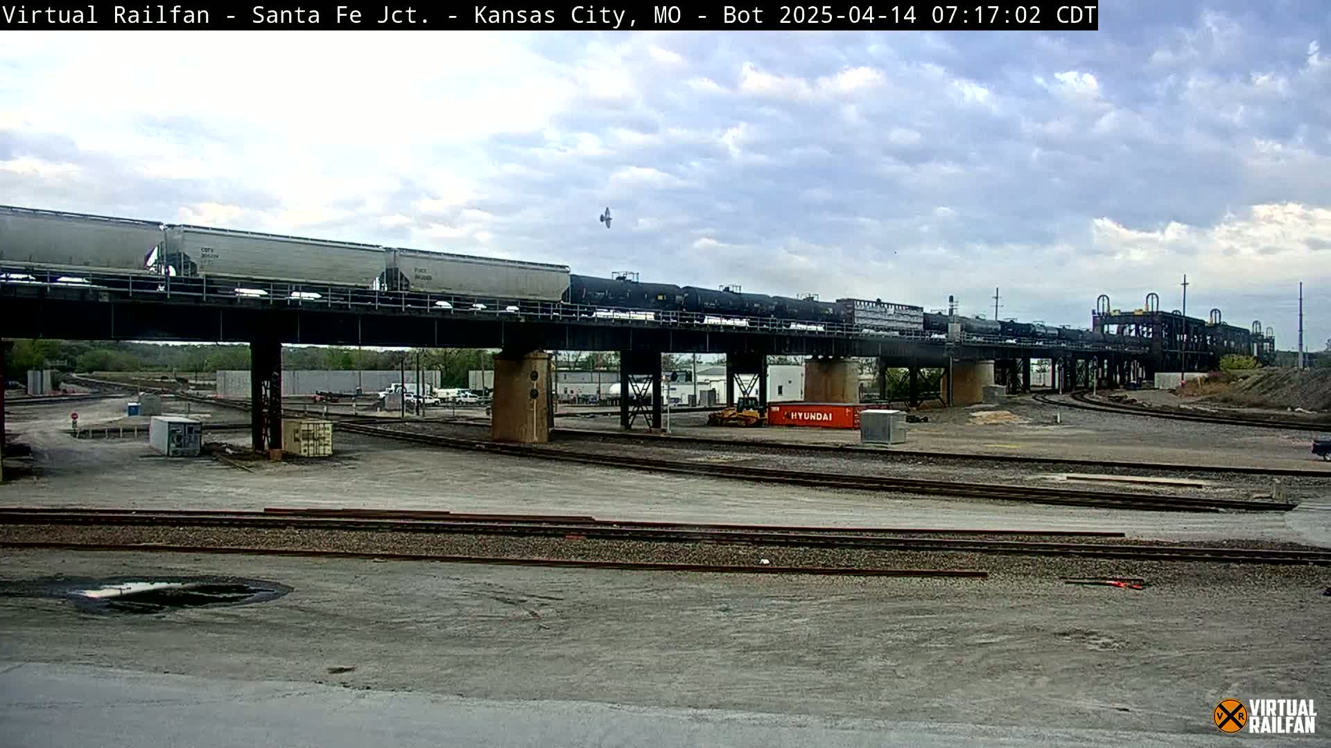 A long freight train crosses a bridge over a rail yard under a partly cloudy sky.