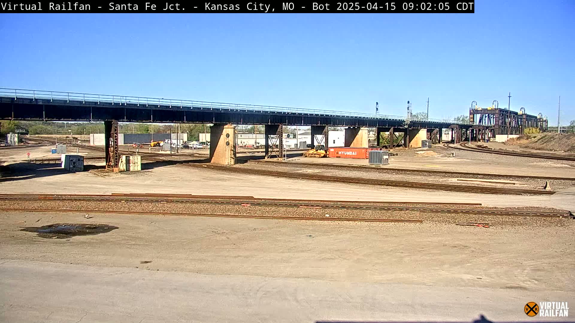 A railway yard with a large bridge under a clear blue sky, showing several train tracks, some construction equipment, and storage containers.