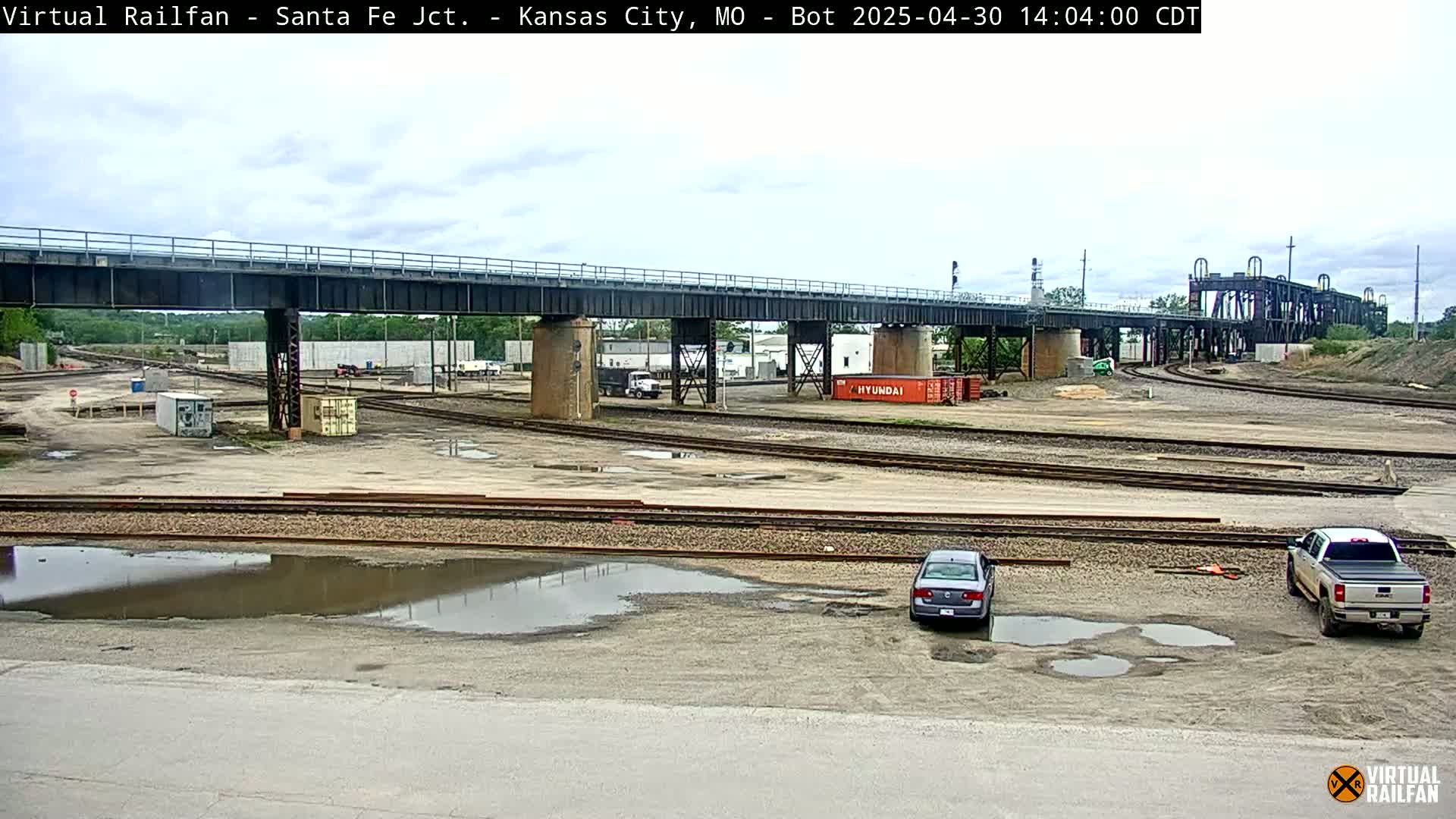 A railway yard with a large bridge overhead under a partly cloudy sky, showing several puddles on the ground and a few cars parked nearby.
