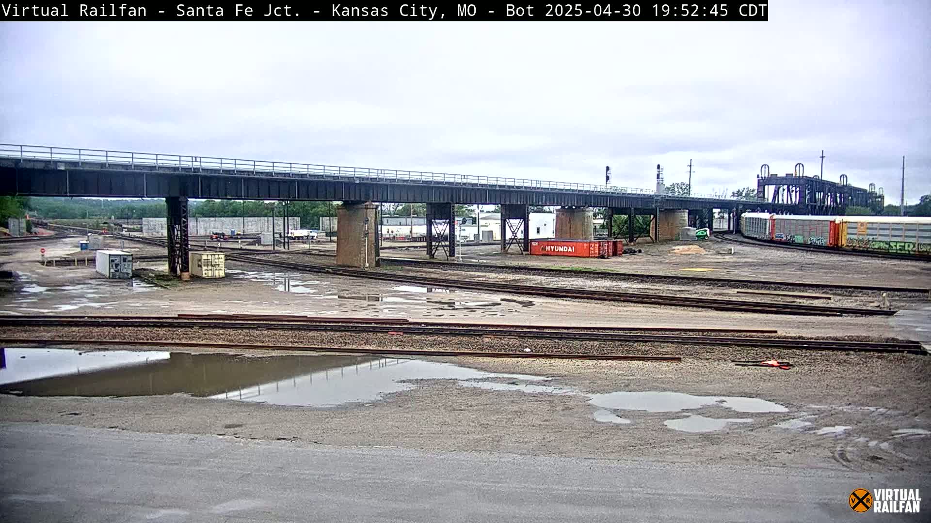 A railway yard with a large bridge overhead, several train cars, and numerous puddles under an overcast sky.