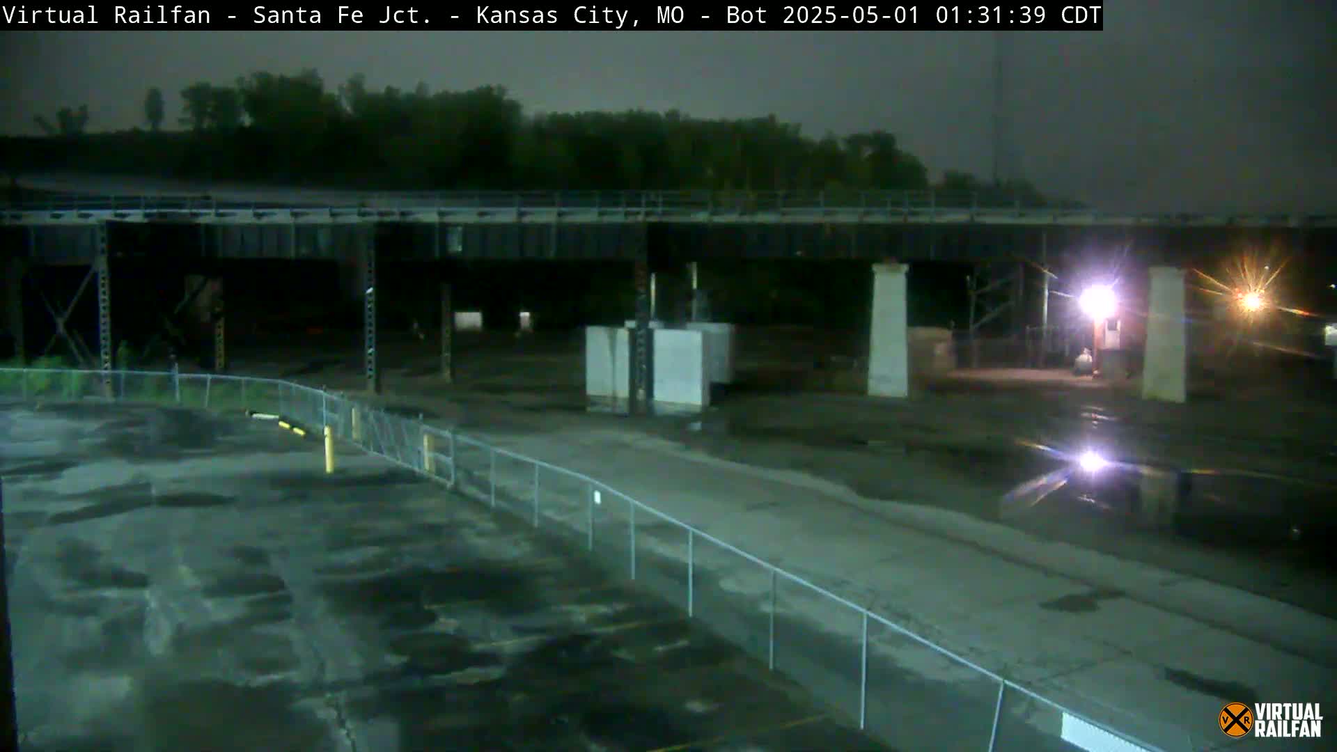 A nighttime view of a mostly empty paved area under a bridge, surrounded by a chain-link fence, with some structures and trees visible in the background under a dark, possibly overcast sky.