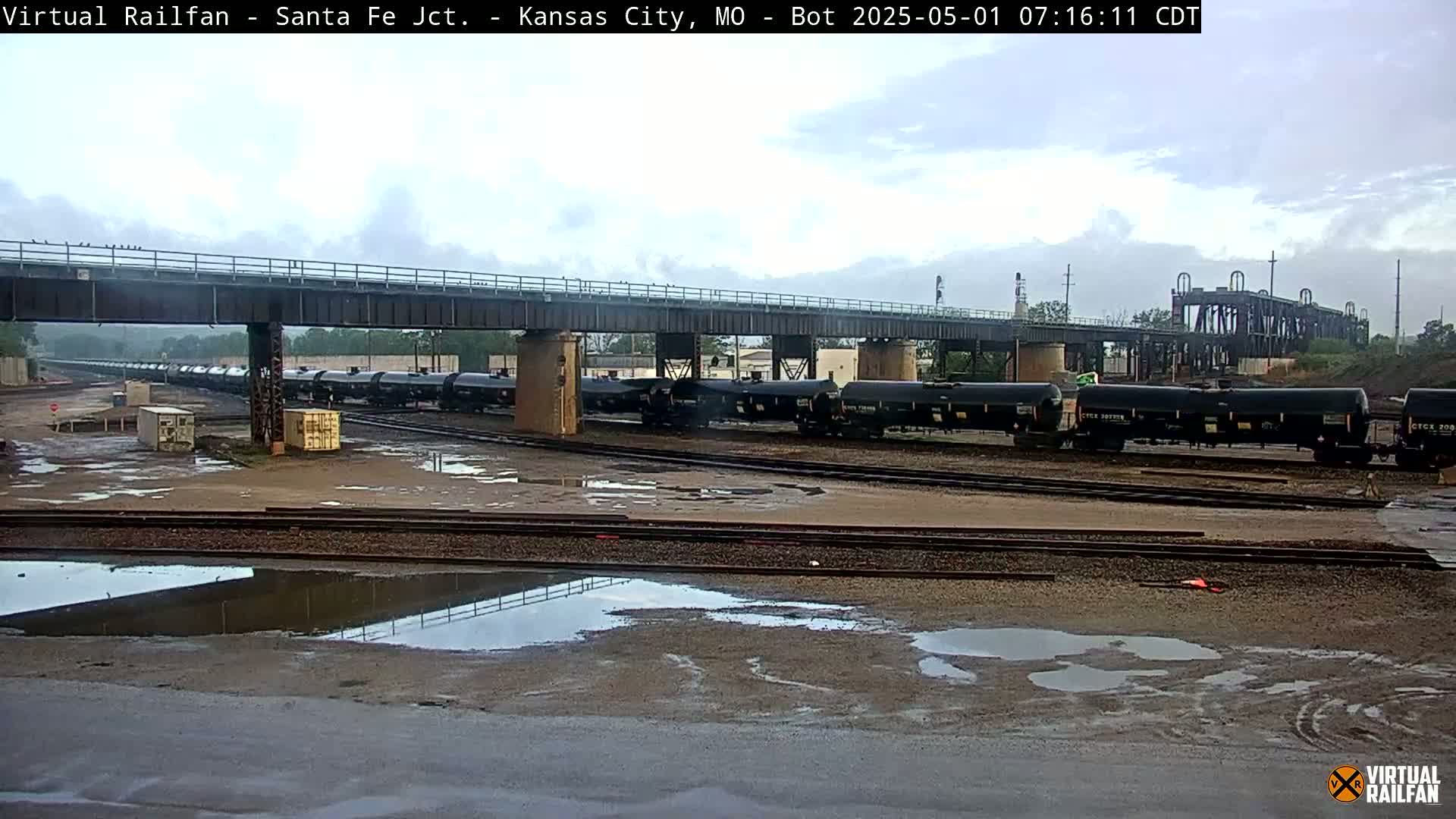 A long train of tanker cars sits on a rail yard under an overcast sky, with puddles of water on the ground.