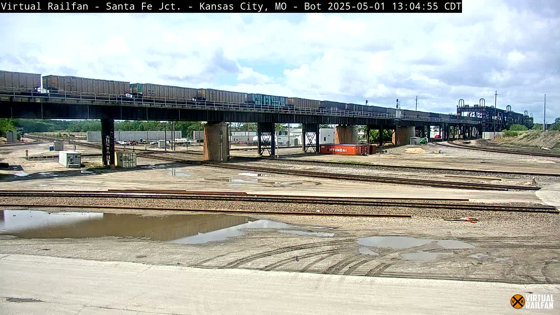 A long freight train crosses a railway bridge over a rail yard under a partly cloudy sky.