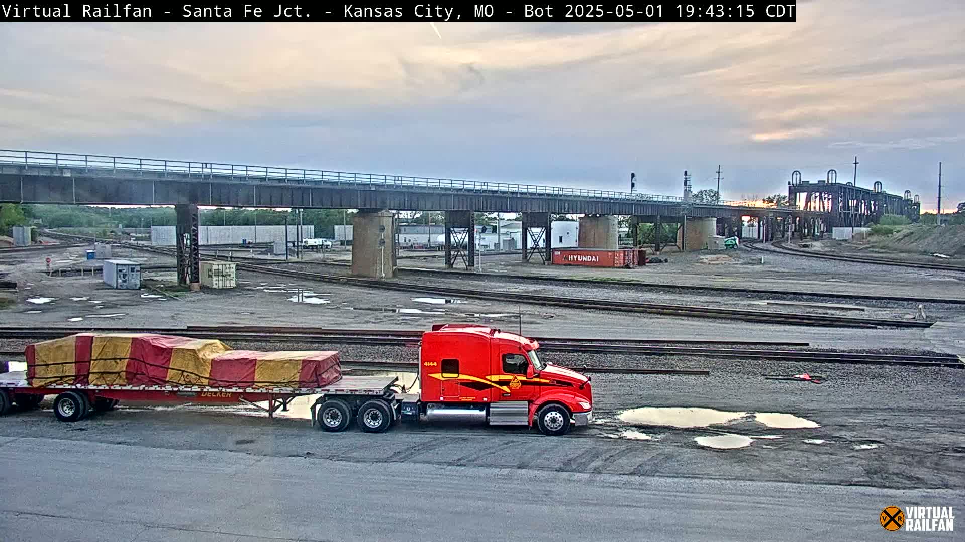 A red semi-truck and trailer sits in a rail yard under an overcast sky, with a bridge visible in the background.