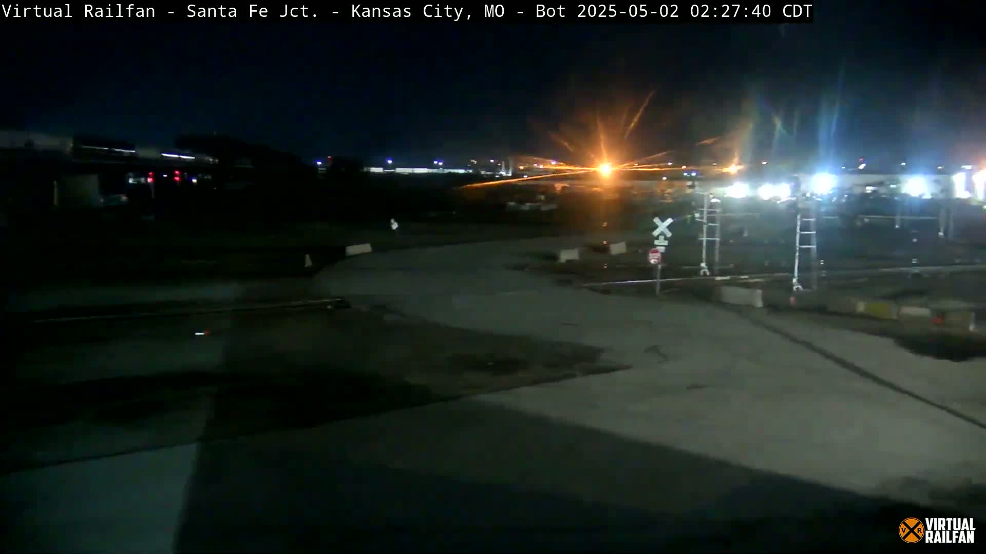 A nighttime view of a railroad crossing with train tracks and a brightly lit area in the background.