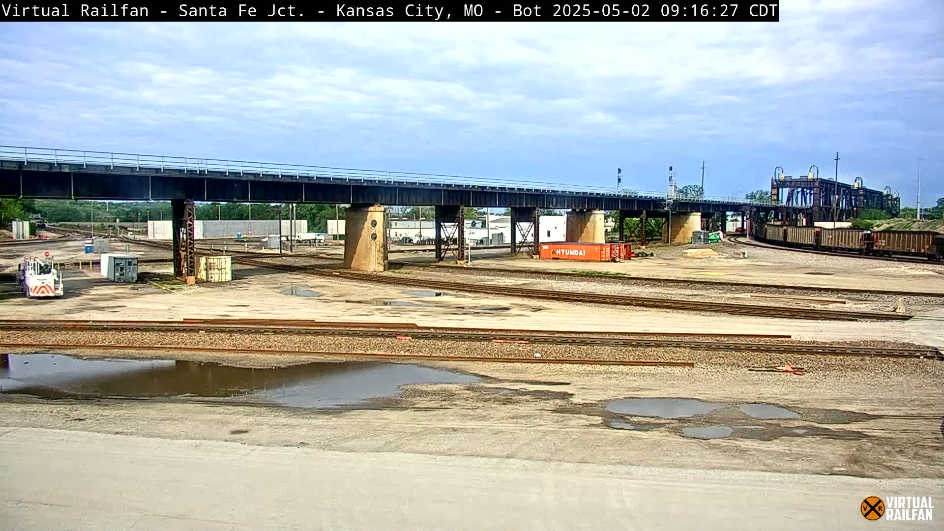 A train yard with several sets of train tracks under a bridge, some puddles of water on the ground, and a partially cloudy sky.