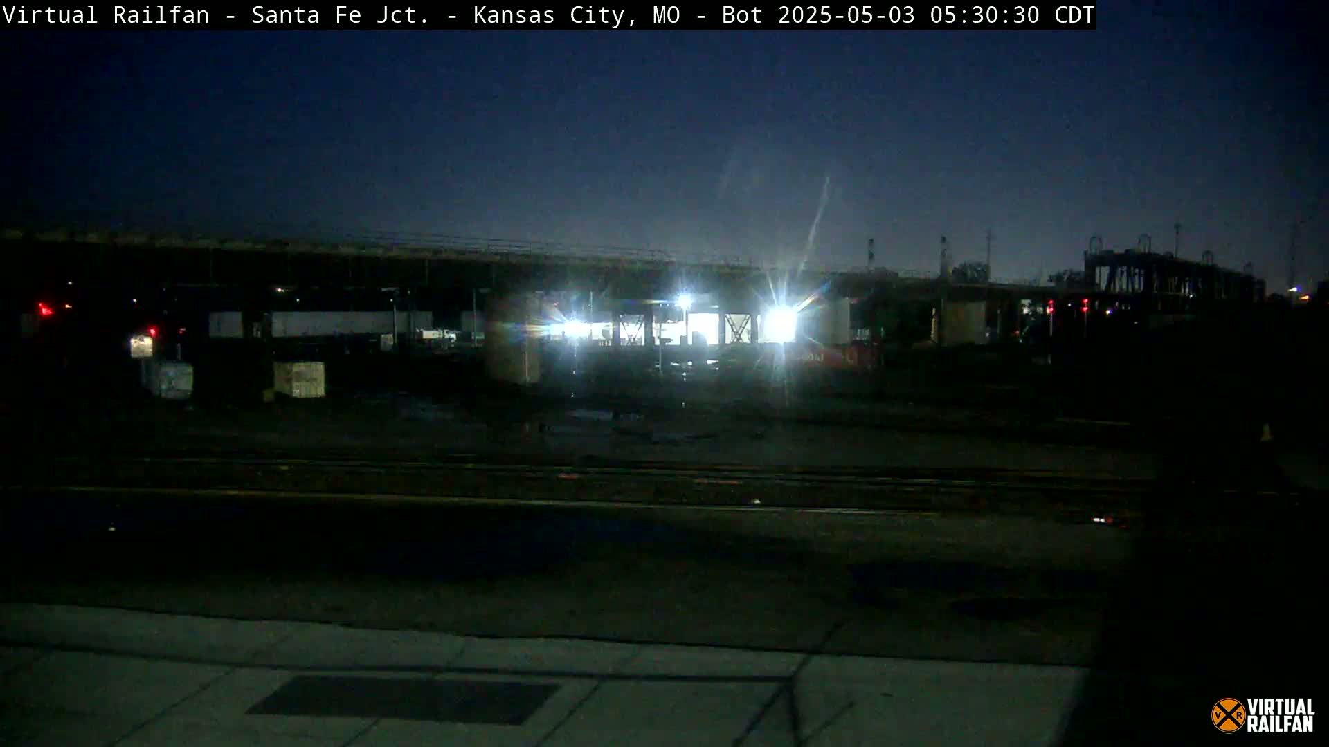 Under a dark night sky, a dimly lit rail yard shows several boxcars and a structure with bright lights.
