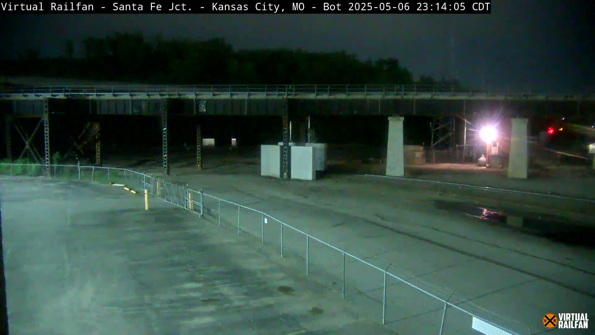 At night, an empty paved area lies beneath a large bridge, enclosed by a chain-link fence.