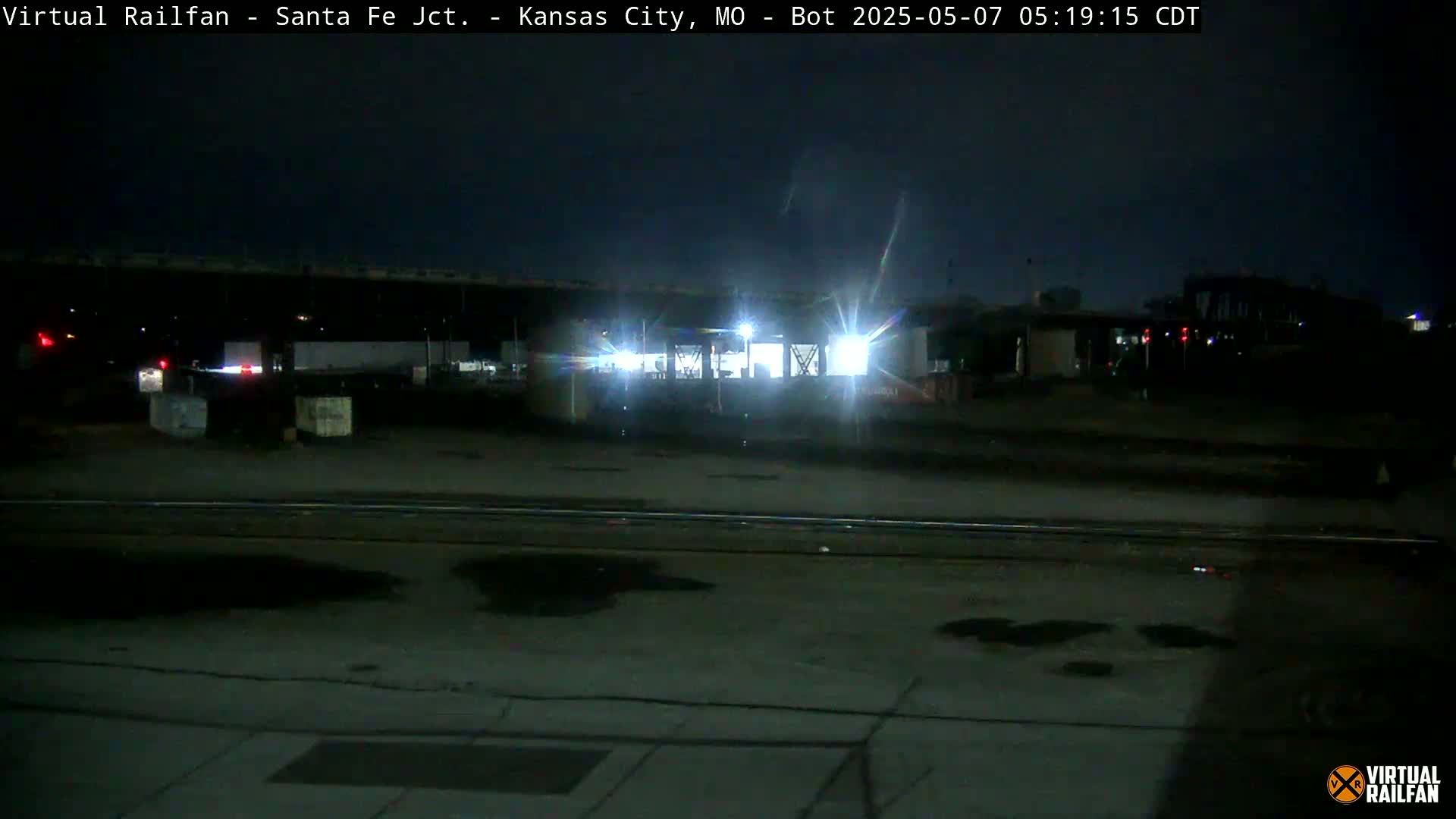 A nighttime view of a dimly lit train yard with tracks and several structures under a dark sky.
