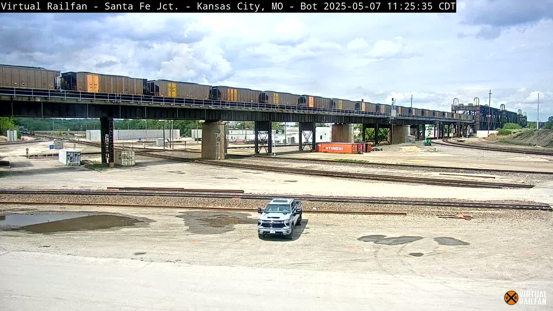 A long freight train crosses a railway bridge over a rail yard on a partly cloudy day, with a light-colored pickup truck parked below.