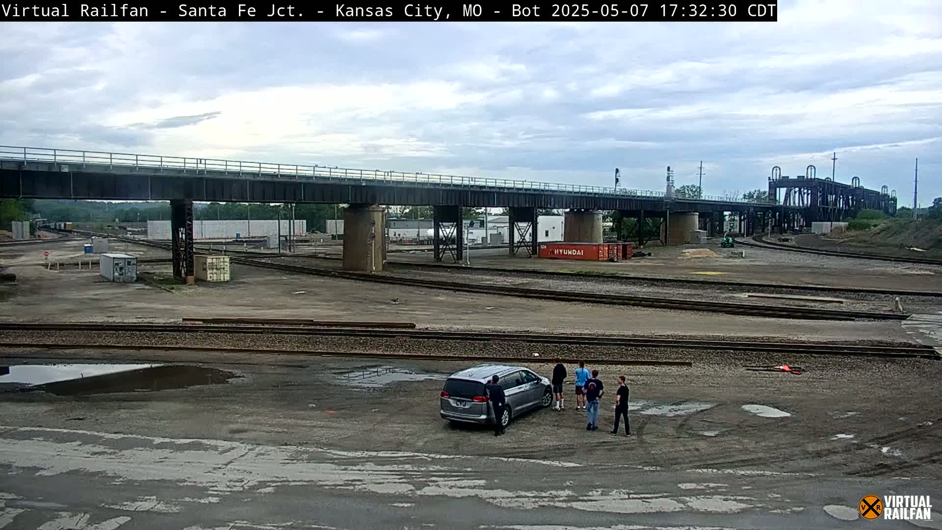 A railway yard with several train tracks, a bridge overhead, and a silver minivan parked near a small group of people under a cloudy sky.