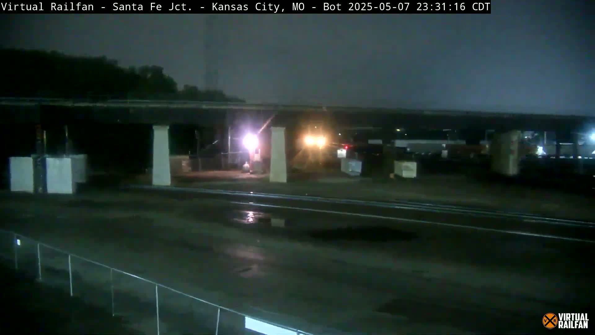 A nighttime view of a wet roadway under a bridge, with some vehicles and structures visible in the distance under overcast skies.