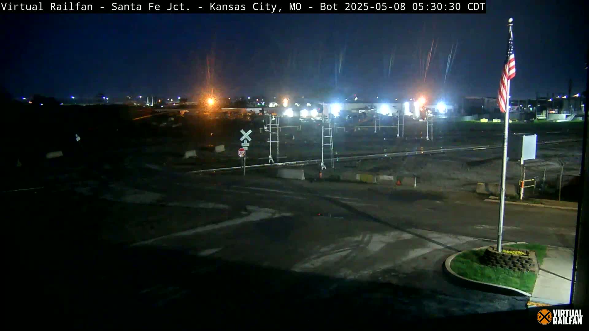 A nighttime view of a mostly empty railroad crossing and surrounding area, with an American flag visible in the foreground.