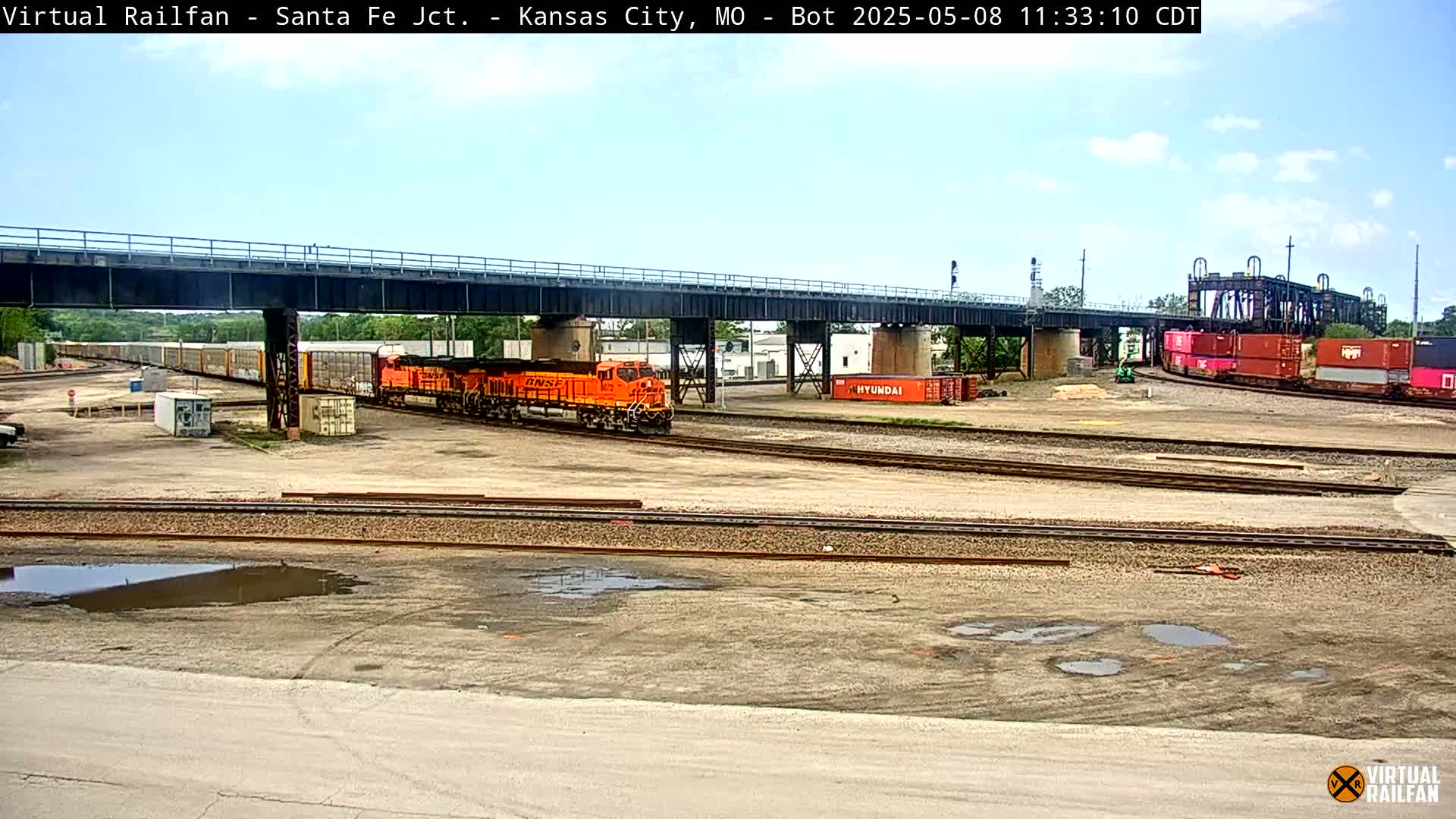An orange locomotive leads a freight train under a bridge on a partly sunny day, with other trains and shipping containers visible in a rail yard.