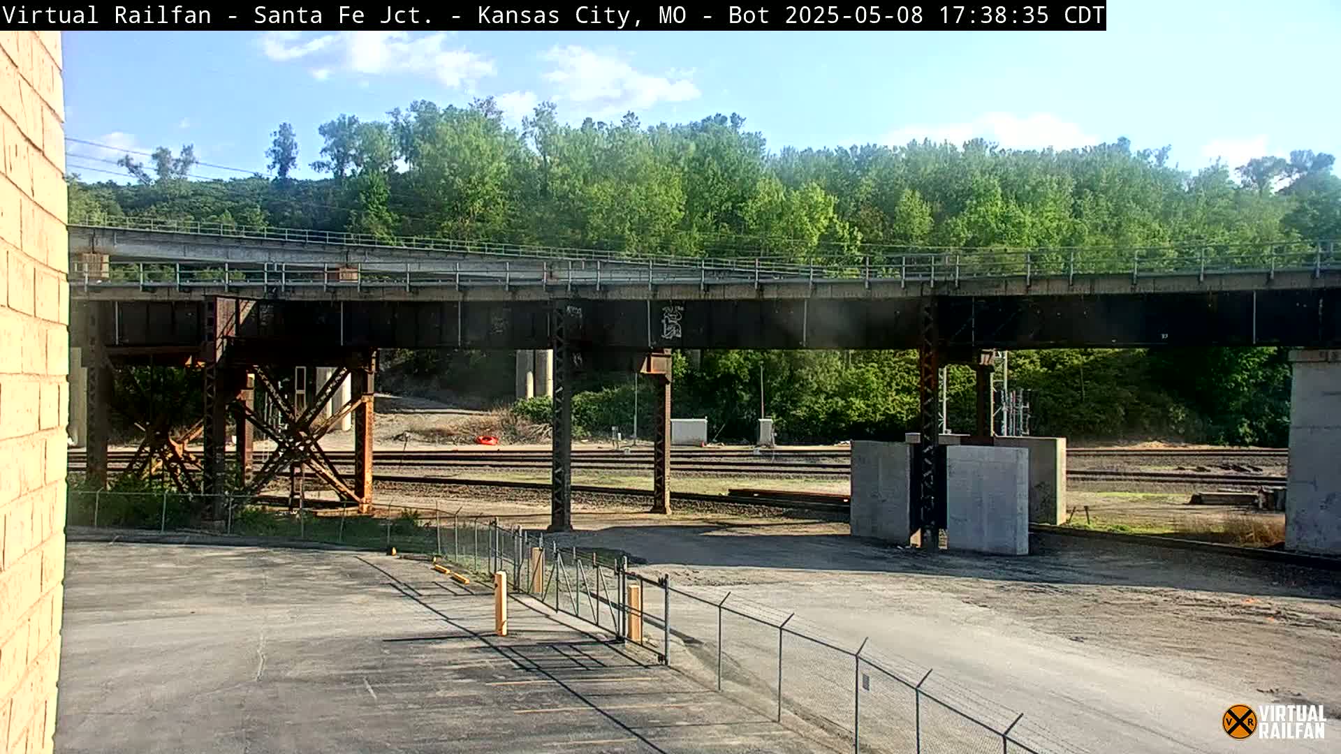A view of railroad tracks and a bridge under a mostly sunny sky, with green trees in the background.