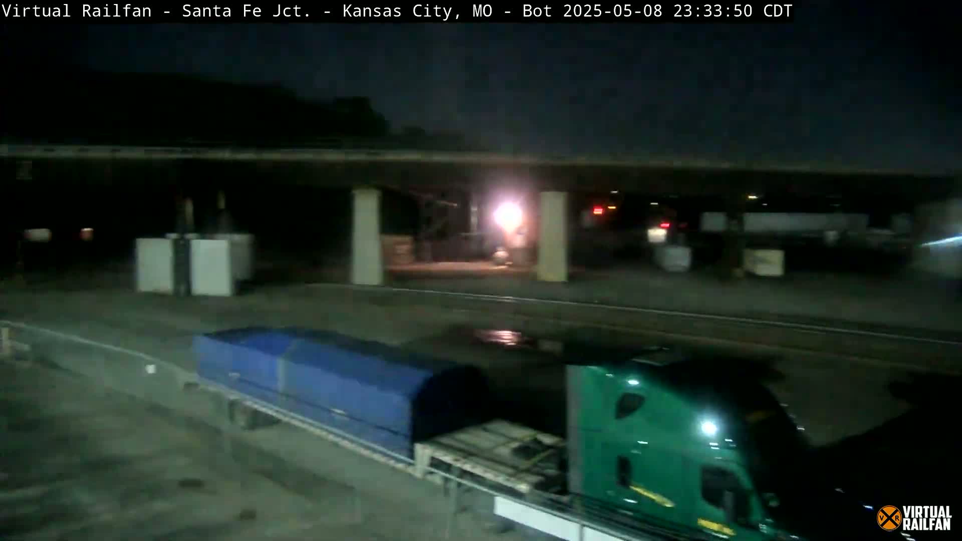 Under a dark night sky, two semi-trucks, one green and one with a blue tarp, are parked near a railway underpass.