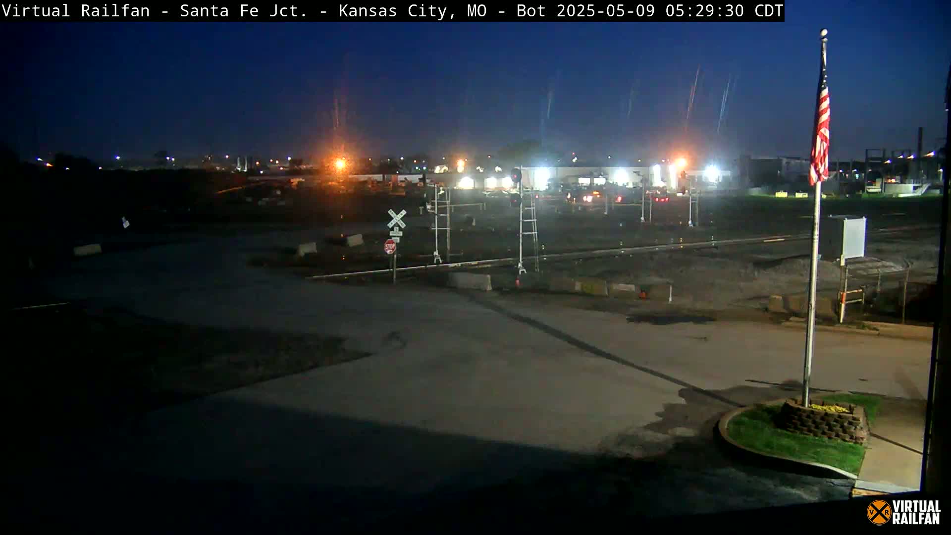 A nighttime view of a railroad crossing with a flagpole and an American flag, under a dark, clear sky.