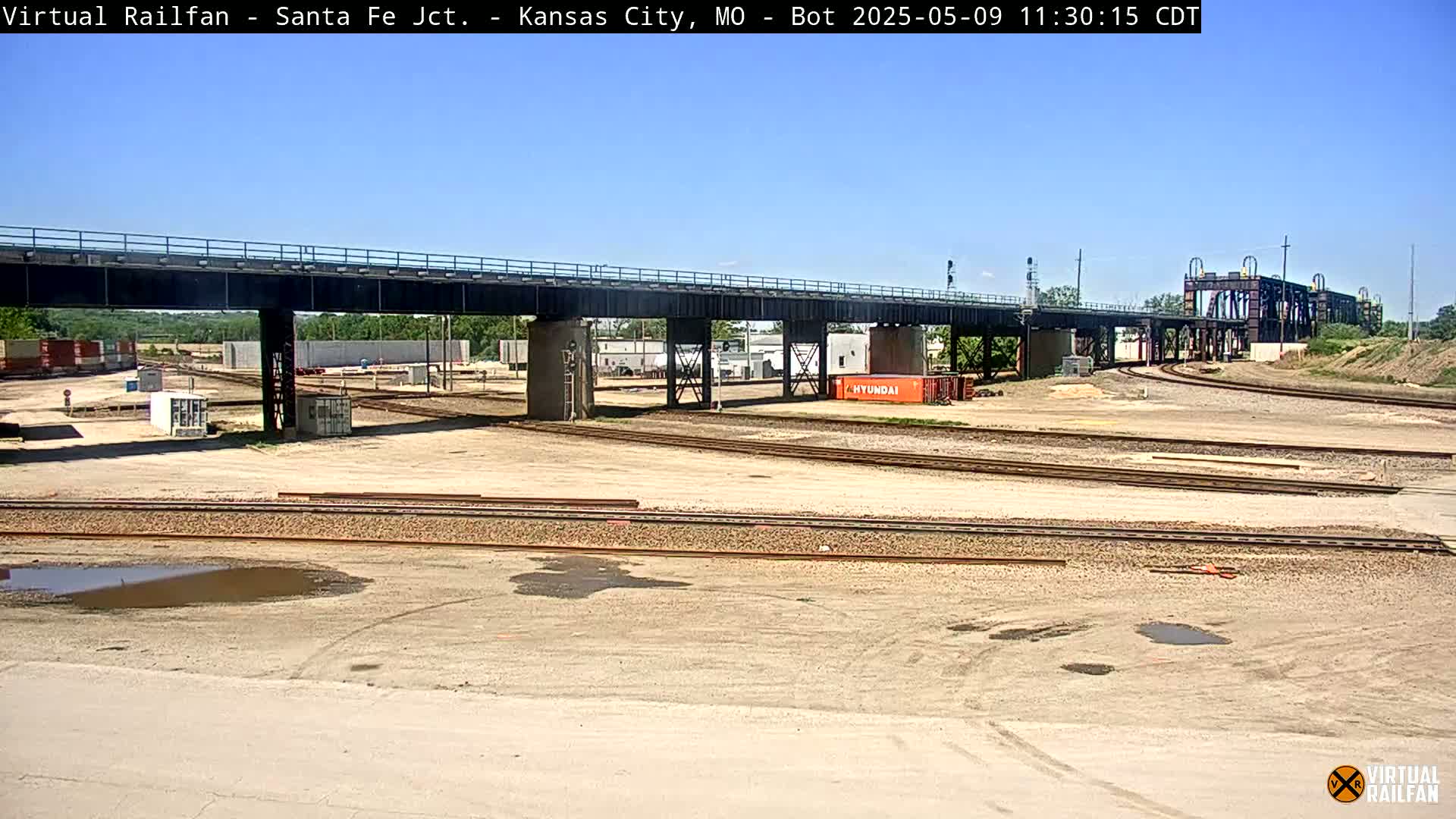 Under a clear blue sky, a railway yard with multiple train tracks stretches beneath a large bridge, and a few shipping containers are visible.