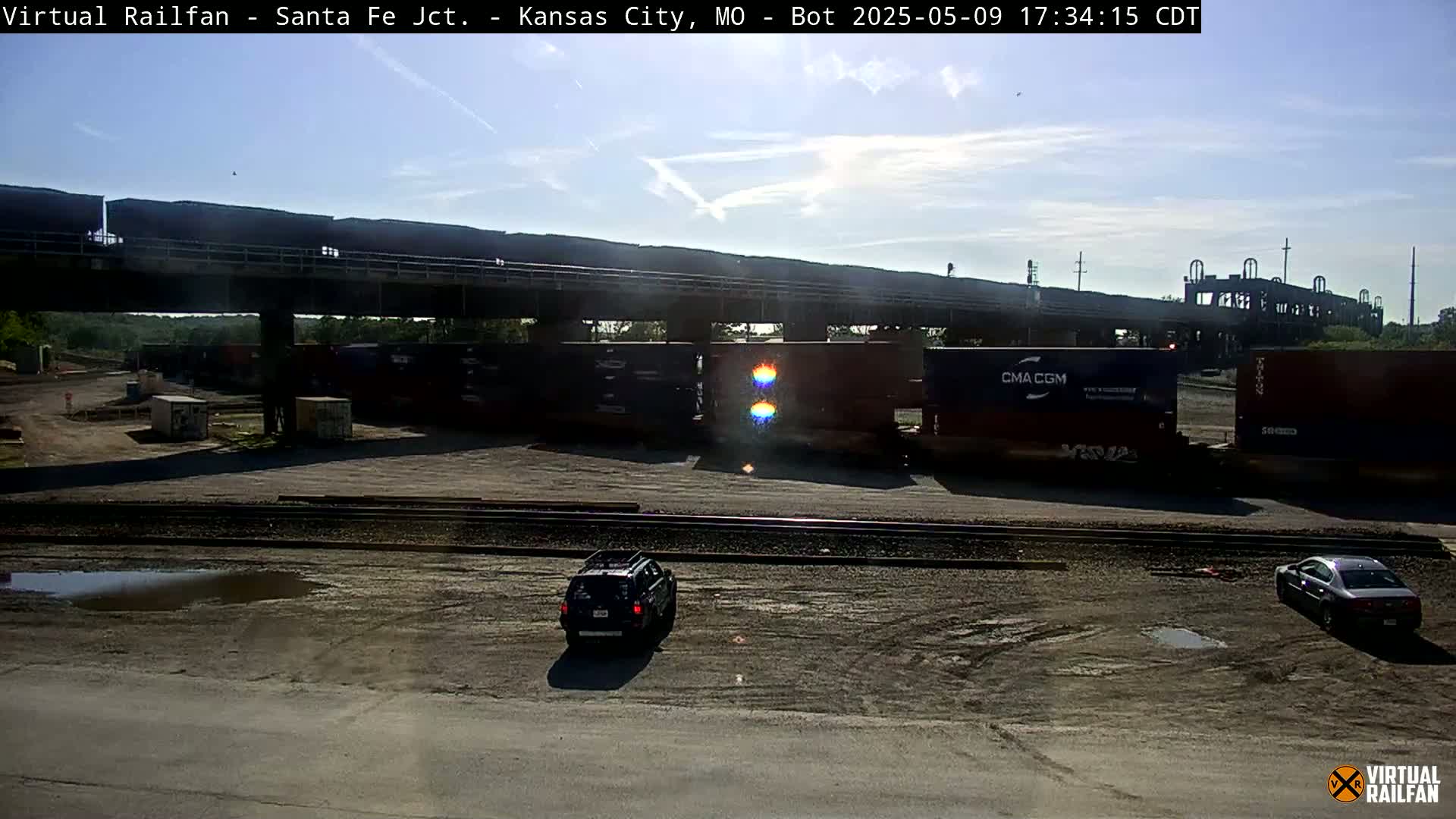 Under a mostly sunny sky, a dark-colored SUV and car are parked near train cars and rail tracks in a rail yard.
