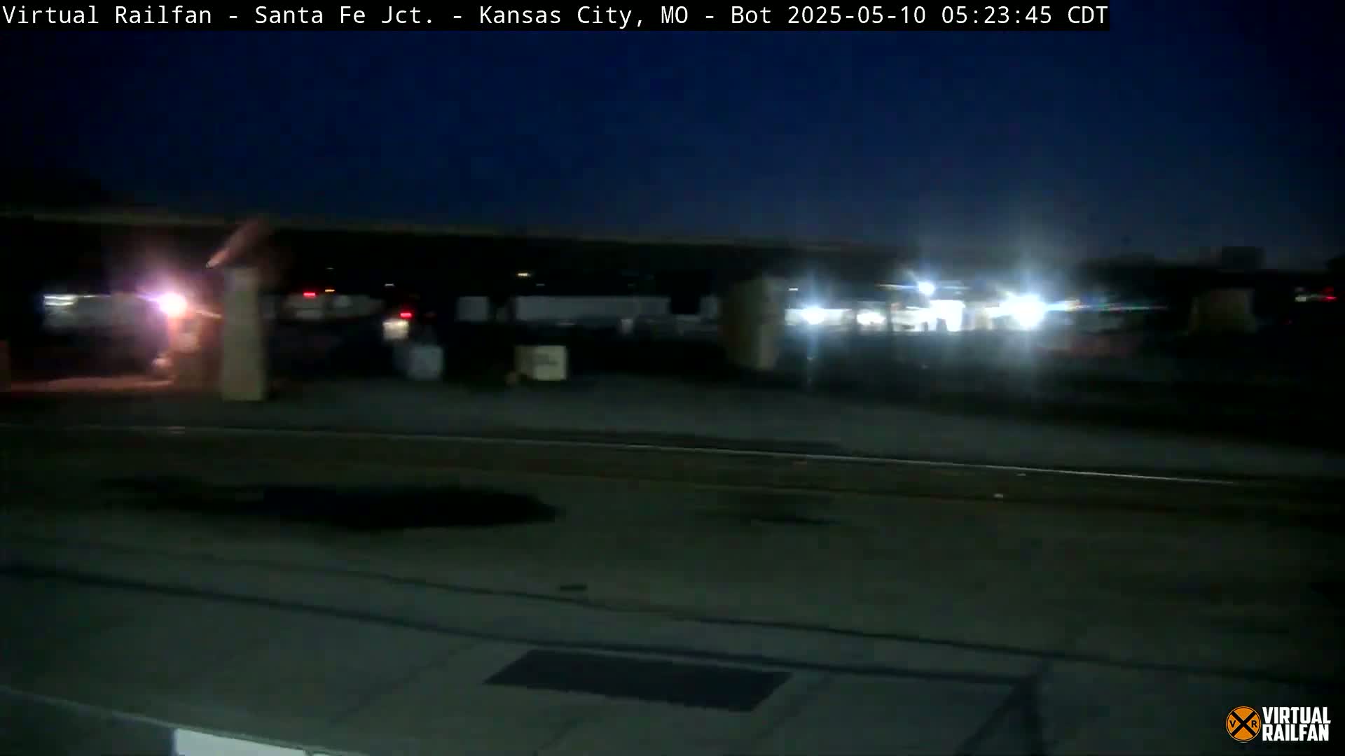 A nighttime view shows a dimly lit area with several vehicles parked under a covered structure near train tracks.