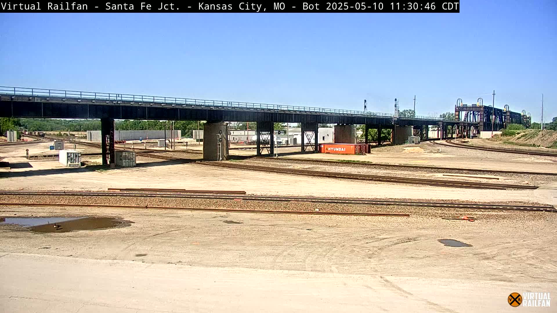 Under a clear blue sky, a railway yard with multiple train tracks and a large bridge stretches across the scene, featuring an orange shipping container.