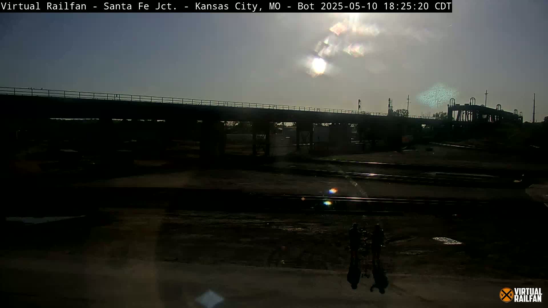 A mostly sunny sky illuminates a railroad yard below a bridge at sunset, with a few people standing near the tracks.