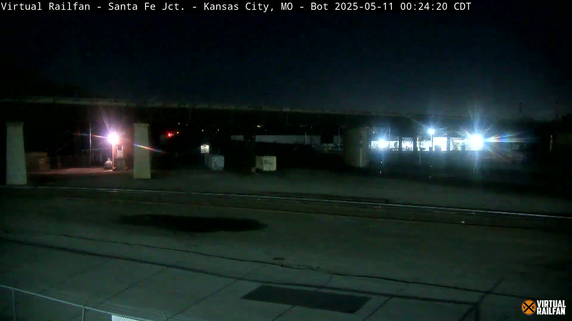 A nighttime, dimly lit view of a rail yard under a bridge, with some structures and vehicles visible.