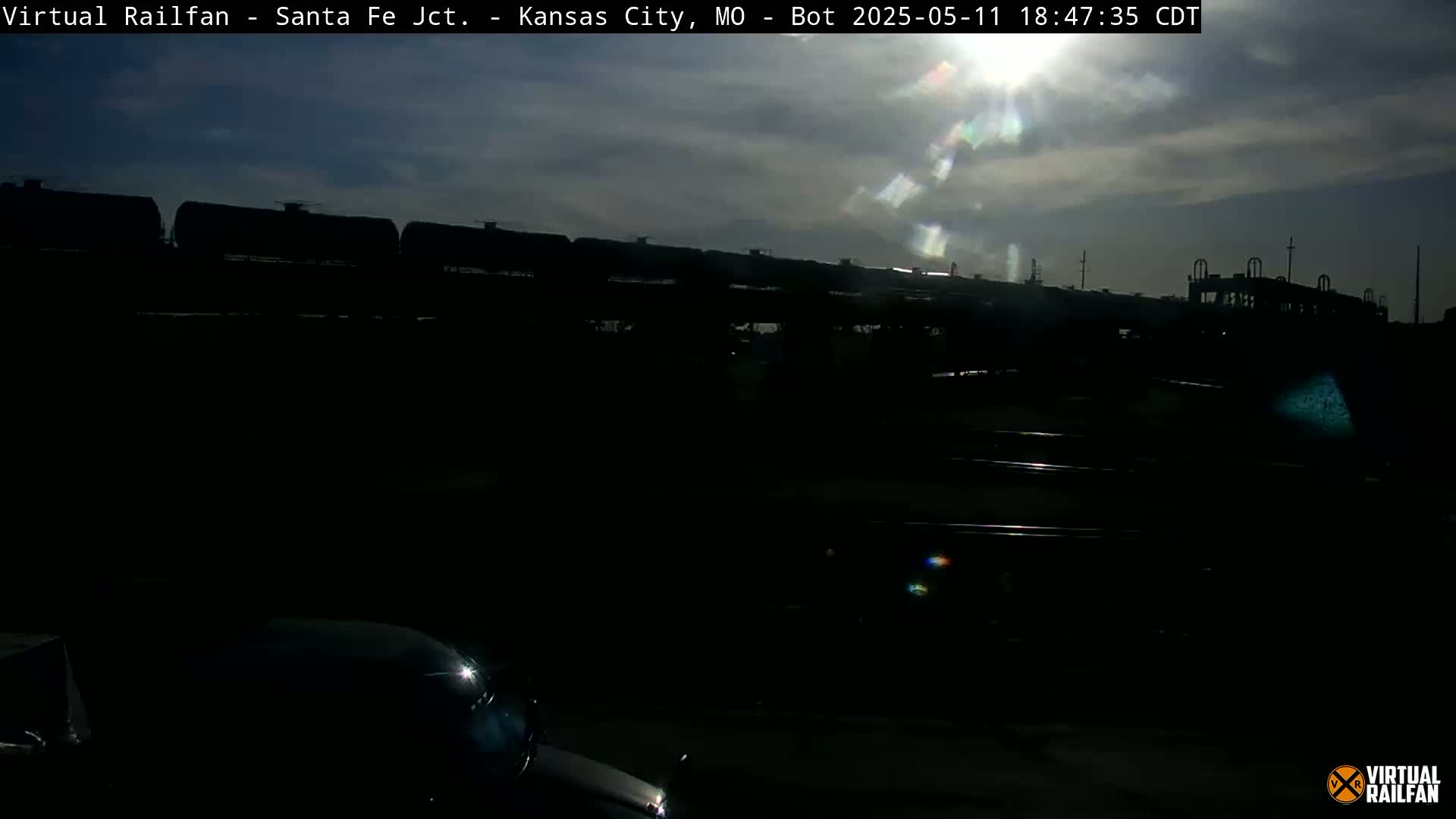 A long freight train of dark tanker cars is visible under a partly sunny sky.