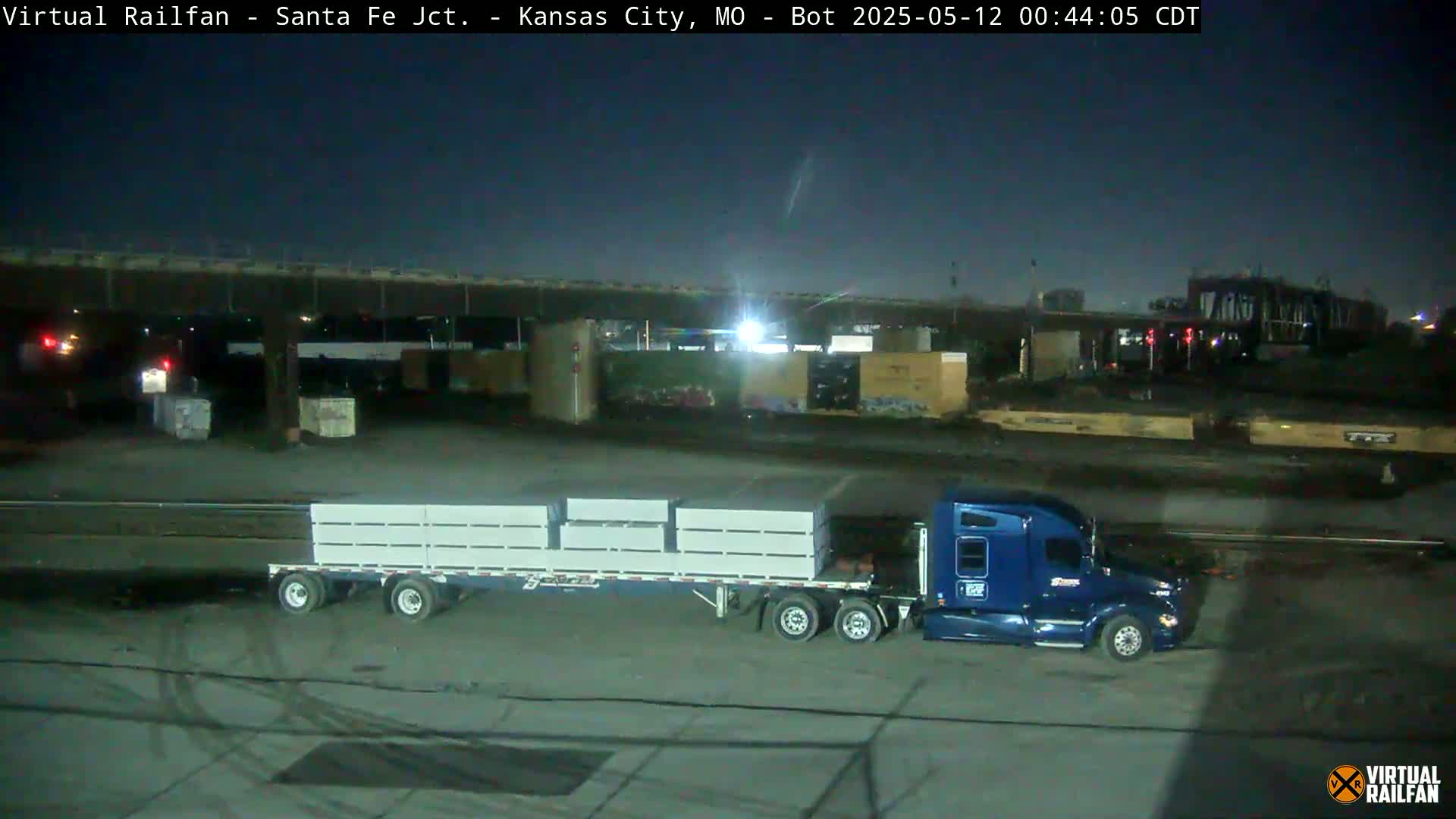 At night, a blue semi-truck hauling a flatbed trailer loaded with white rectangular containers sits in a large, empty lot near railroad tracks and under a bridge.