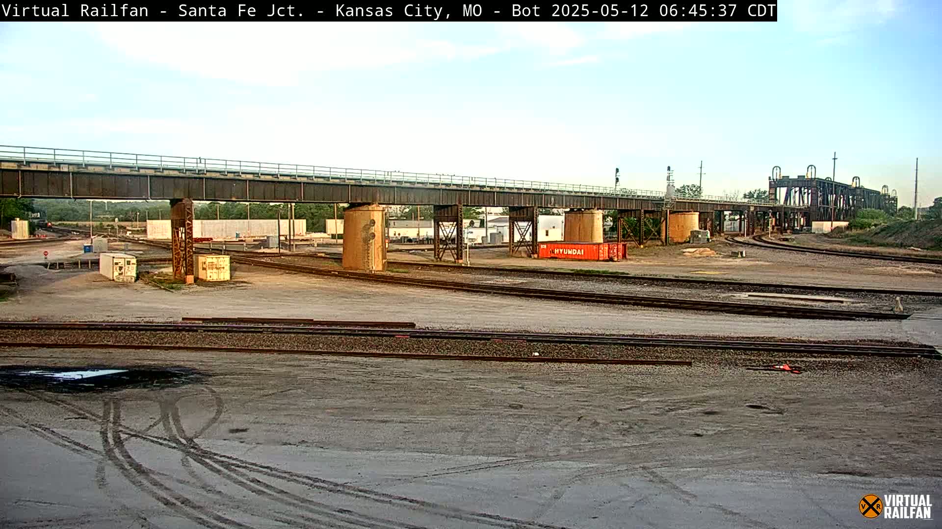 A train yard with railway tracks, a large bridge, and several shipping containers under a partly cloudy sky.