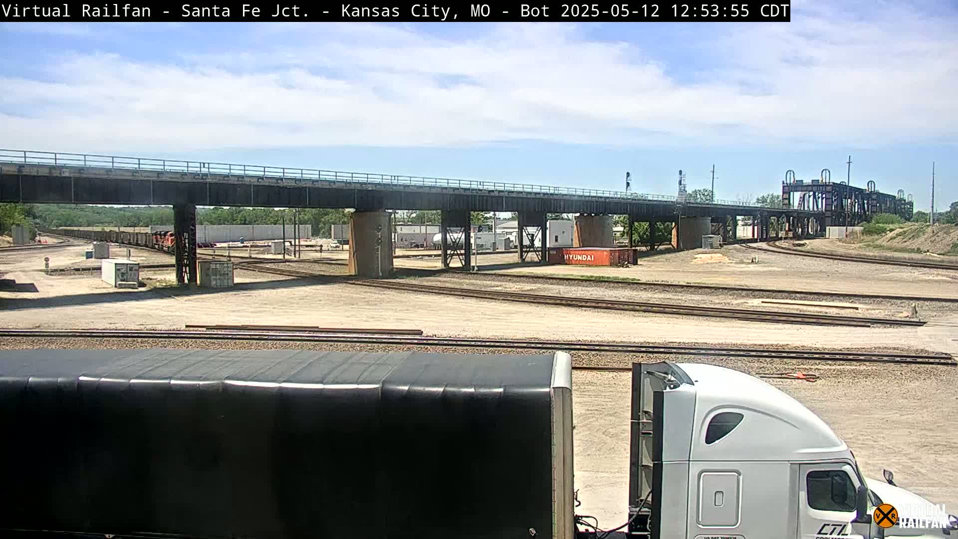 A sunny day reveals a rail yard with a train, several shipping containers, and a large bridge, all viewed from behind a semi-truck.