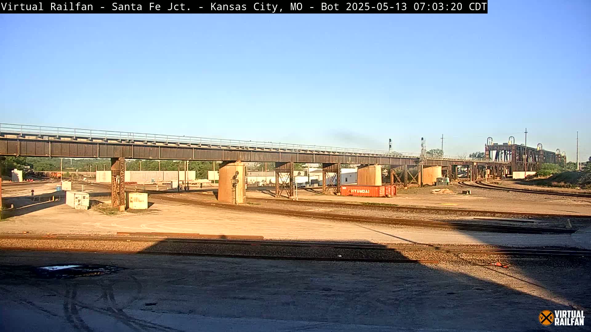 A railway yard with a large metal bridge under a clear blue sky.
