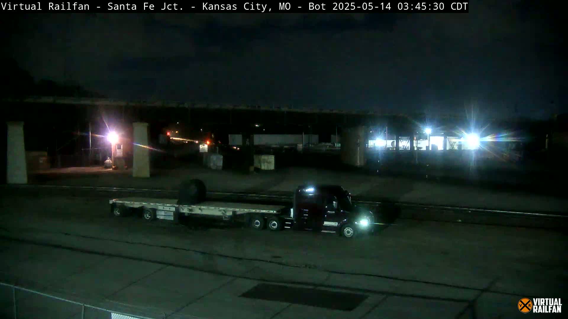 At night, a flatbed semi-truck is parked under a bridge near a rail yard.
