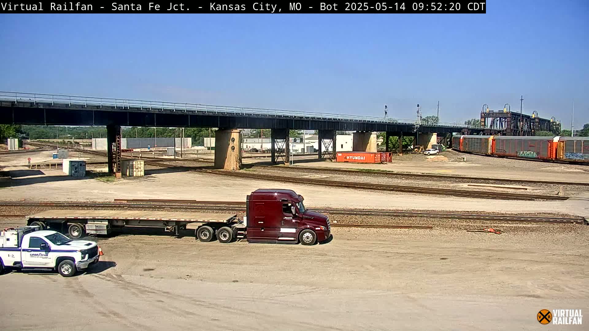 A maroon semi-truck and a white pickup truck are parked near railroad tracks under a bridge on a sunny day, with a freight train visible in the background.