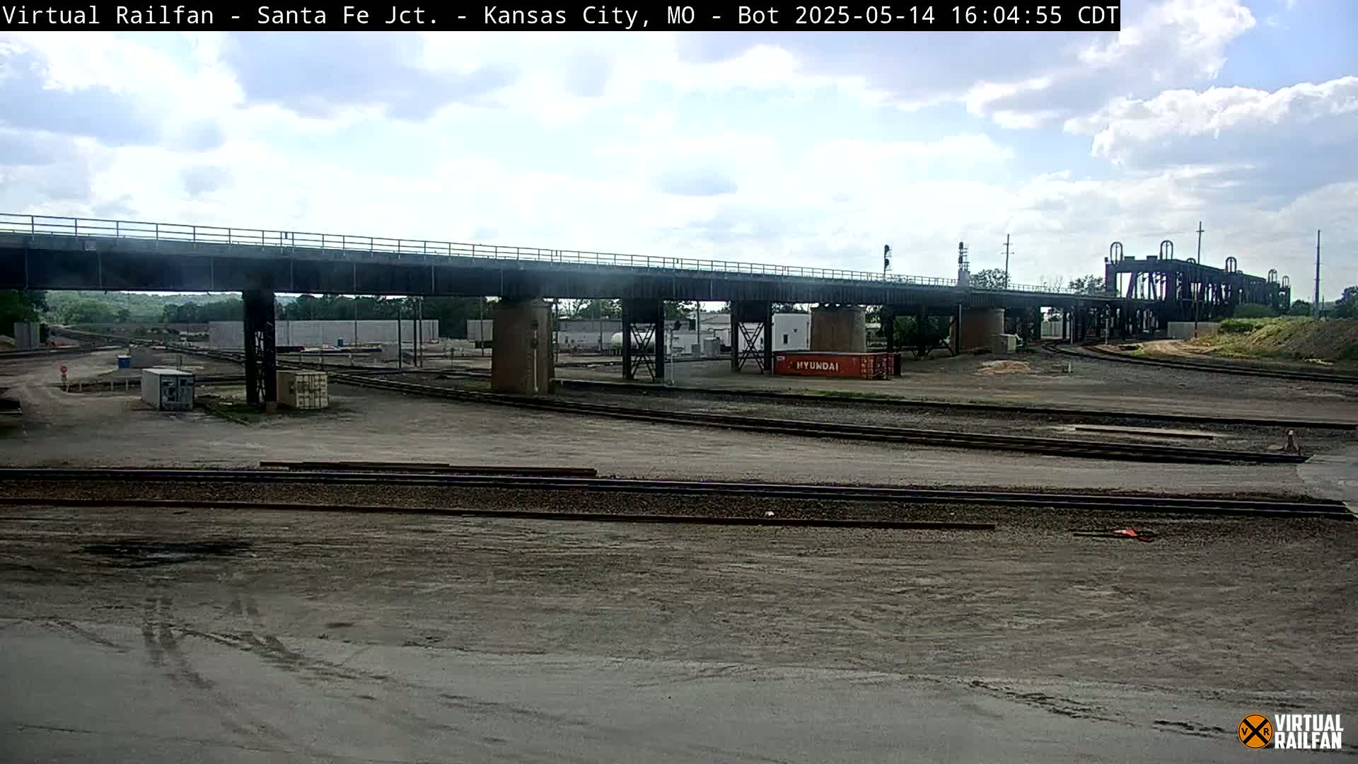 A railway yard under a bridge on a partly cloudy day, with railroad tracks and a few shipping containers visible.