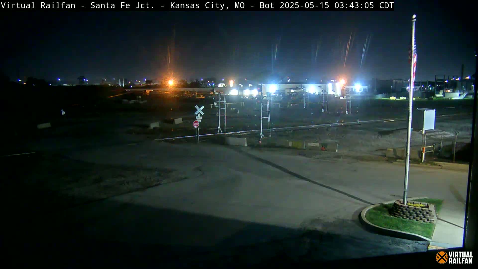 A nighttime view of a railroad crossing with a stop sign, under a clear sky, showing an empty road and some distant lights.