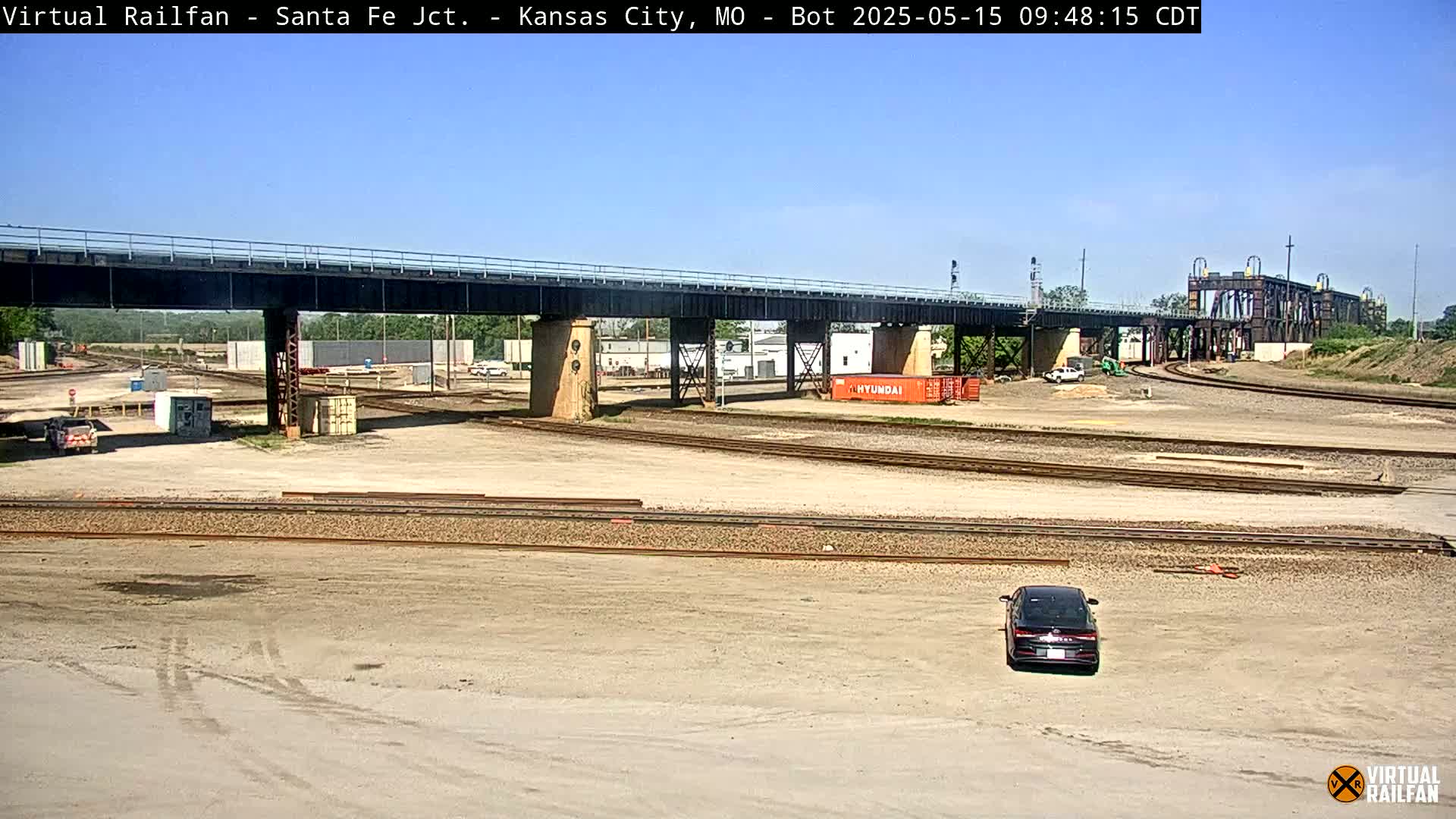 A black car sits in a large, mostly empty rail yard under a bridge on a sunny day.