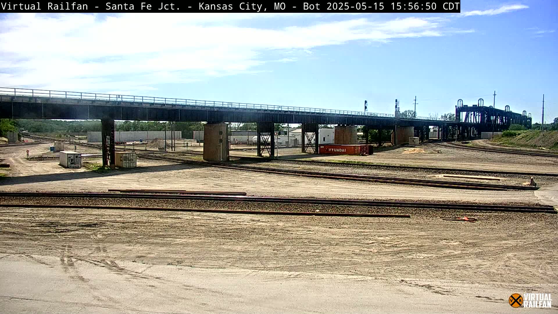 A railway yard under a bridge on a sunny day, with railway tracks and some shipping containers visible.