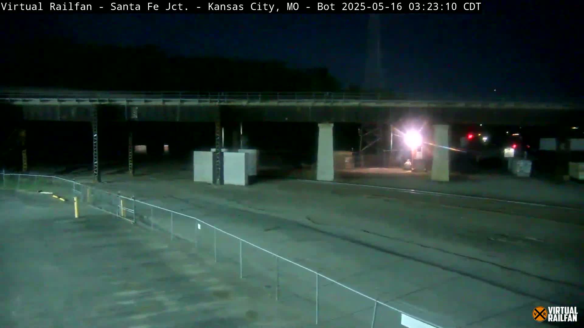 A nighttime view of a mostly empty paved area under a bridge, partially enclosed by a chain-link fence.