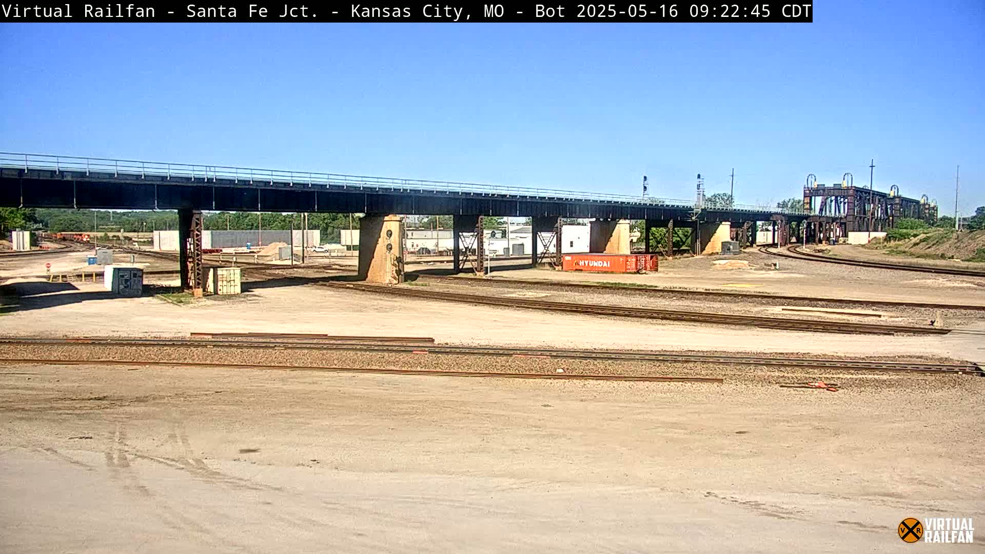 A railroad yard with a large bridge overhead under a clear blue sky.