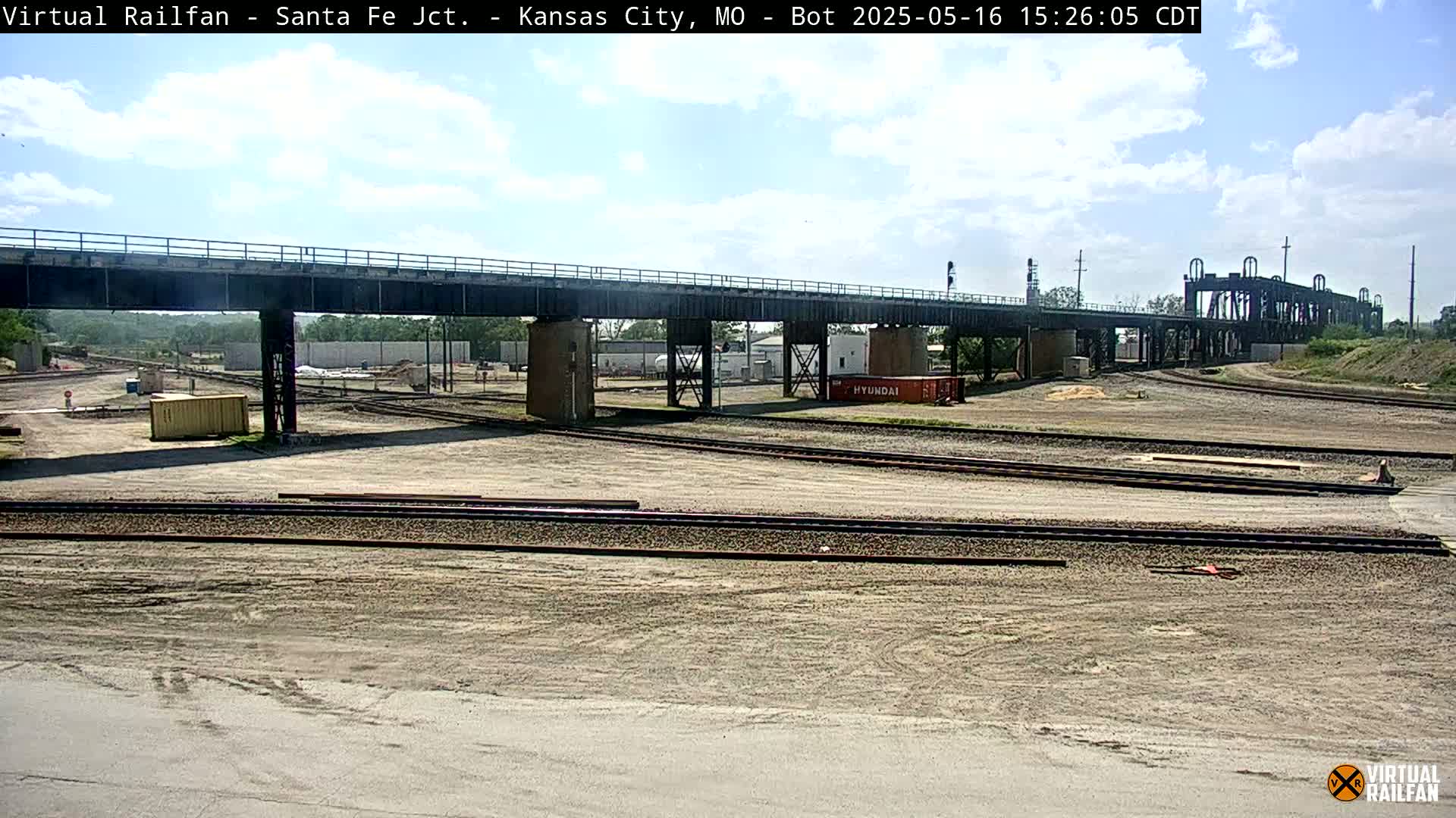 A railway yard with a large bridge under mostly sunny skies, featuring multiple sets of train tracks and some construction materials.