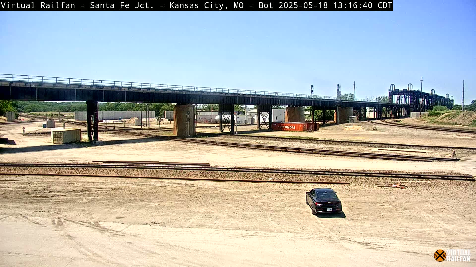 A black car sits in a large, mostly empty rail yard under a sunny sky, with a bridge and several train tracks visible.
